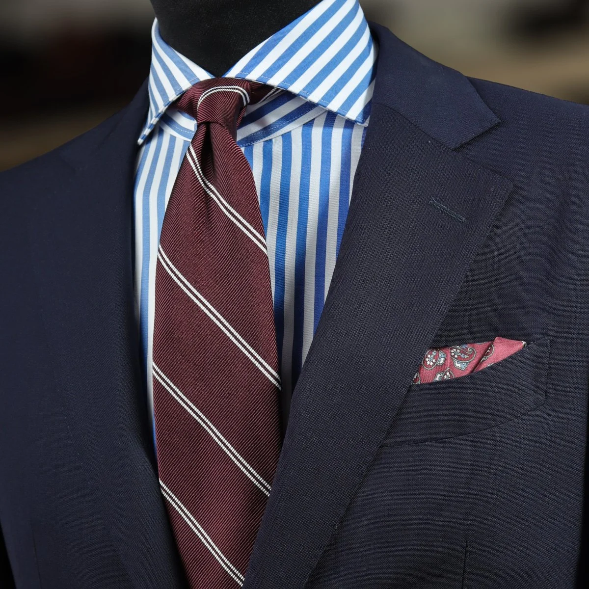 Tamsiai raudonas dryžuotas kaklaraištis, dryžuoti marškiniai ir mėlynas kostiumas